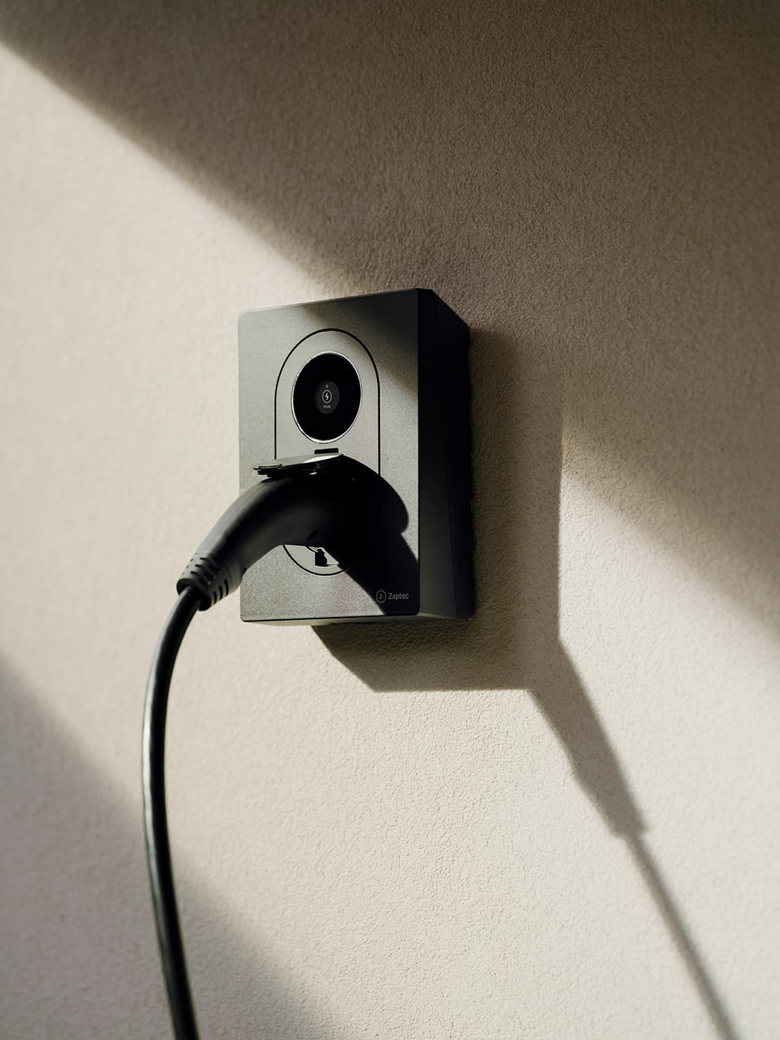 Nahaufnahme Zaptec Go 2 Ladestation eingesteckt an weißer Wand, starker Schatten und Sonnenlicht auf der Oberfläche.