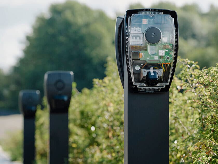 Close-up of Zaptec Pro charger mounted on column in parking lot, more chargers on columns in background, greenery around.