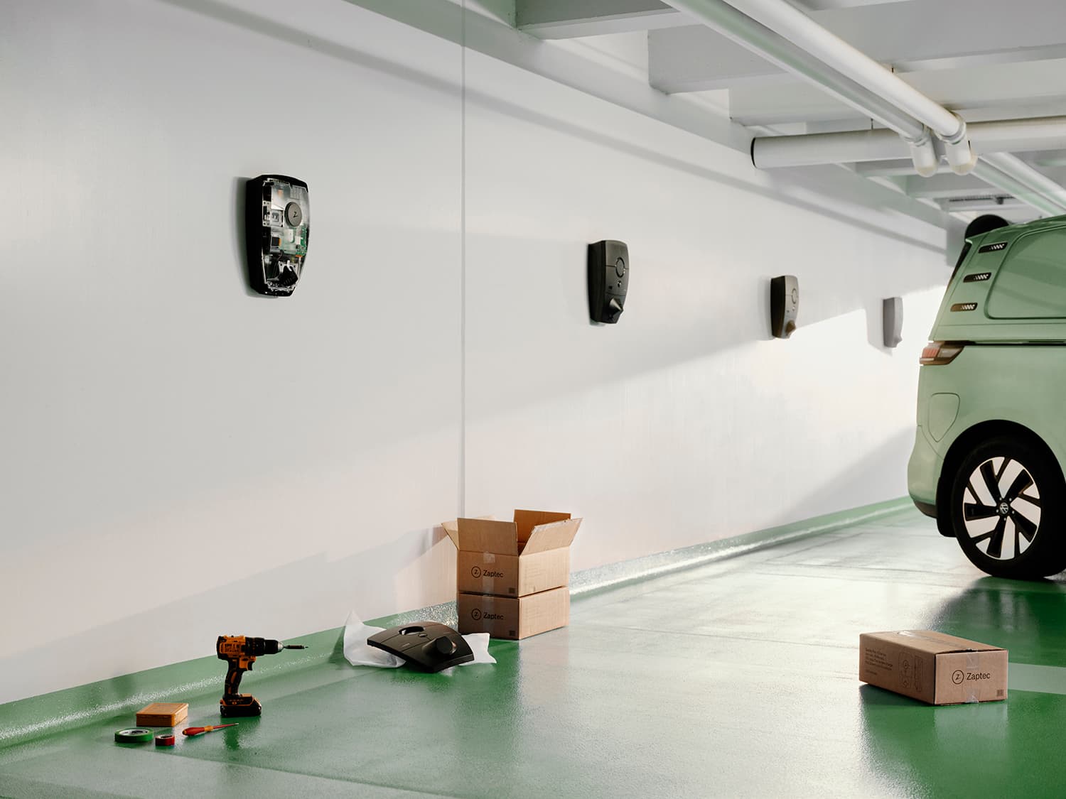 Multiple Zaptec Pro chargers mounted on white wall in parking garage, green van, drill, cardboard boxes, and charger cover on floor.