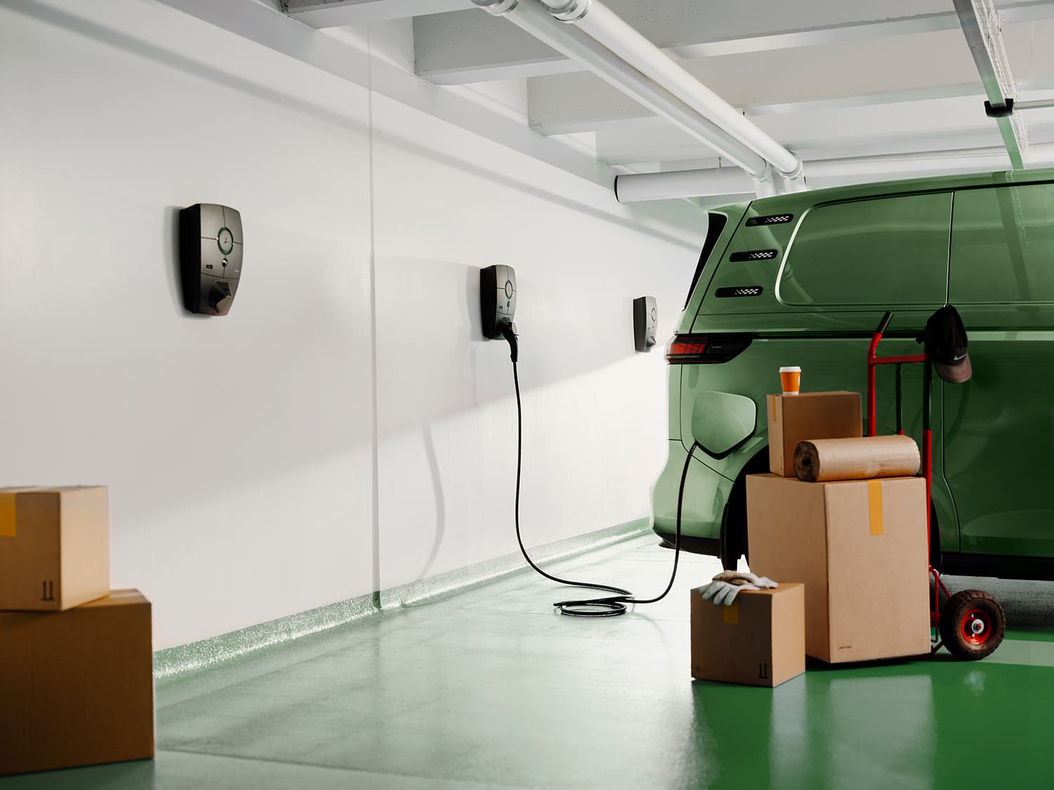 Fourgon vert en charge avec borne Zaptec Pro mur blanc en parking, cartons empilés, gants et gobelet à café au premier plan.