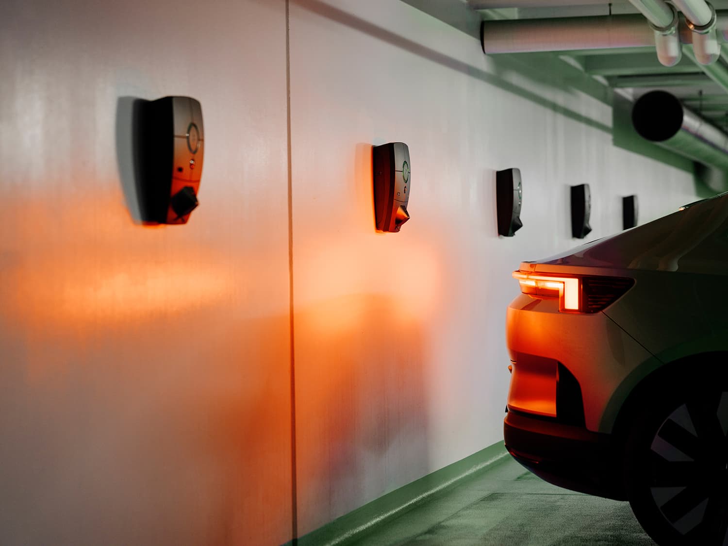 Meerdere Zaptec Pro laders op witte muur in parkeergarage, rood achterlicht van groene auto weerspiegelt op muur, dramatische verlichting.