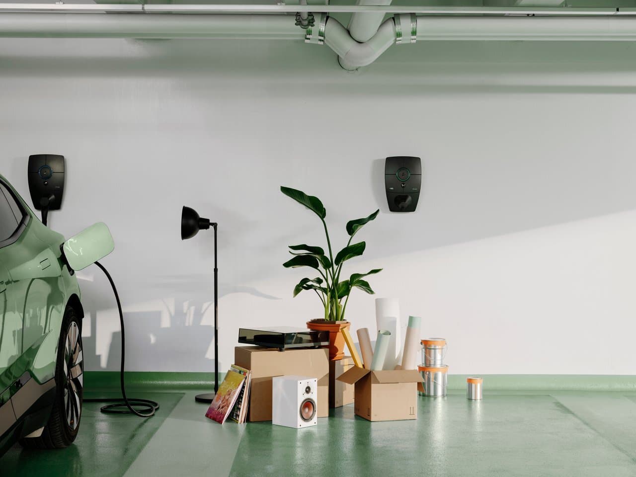 Green car charging with Zaptec Pro charger on white wall in parking garage, boxes, houseplant, lamp, paint cans, and vinyl records, moving or renovation scene.