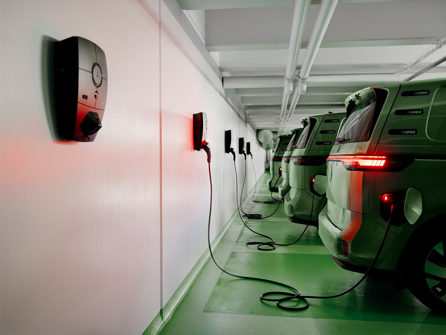 Rangée de fourgons verts en charge avec bornes Zaptec Pro mur blanc en parking, feux arrière rouges allumés.