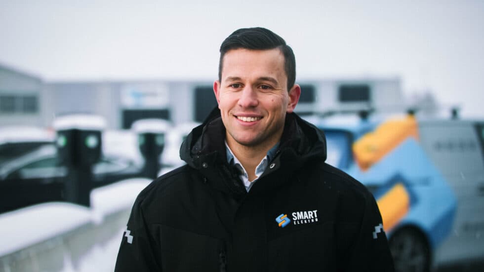 En mann iført en svart jakke med Smart Elektro-logoen smiler foran en vinterlig bakgrunn med elbilladere, snødekte biler og en firmabil i bakgrunnen.