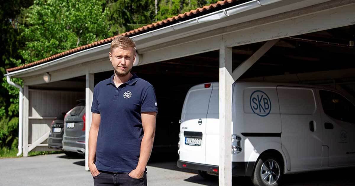 en man med blå pike och svarta byxor står framför en carport med händerna i fickorna i carporten står en vit firmabil