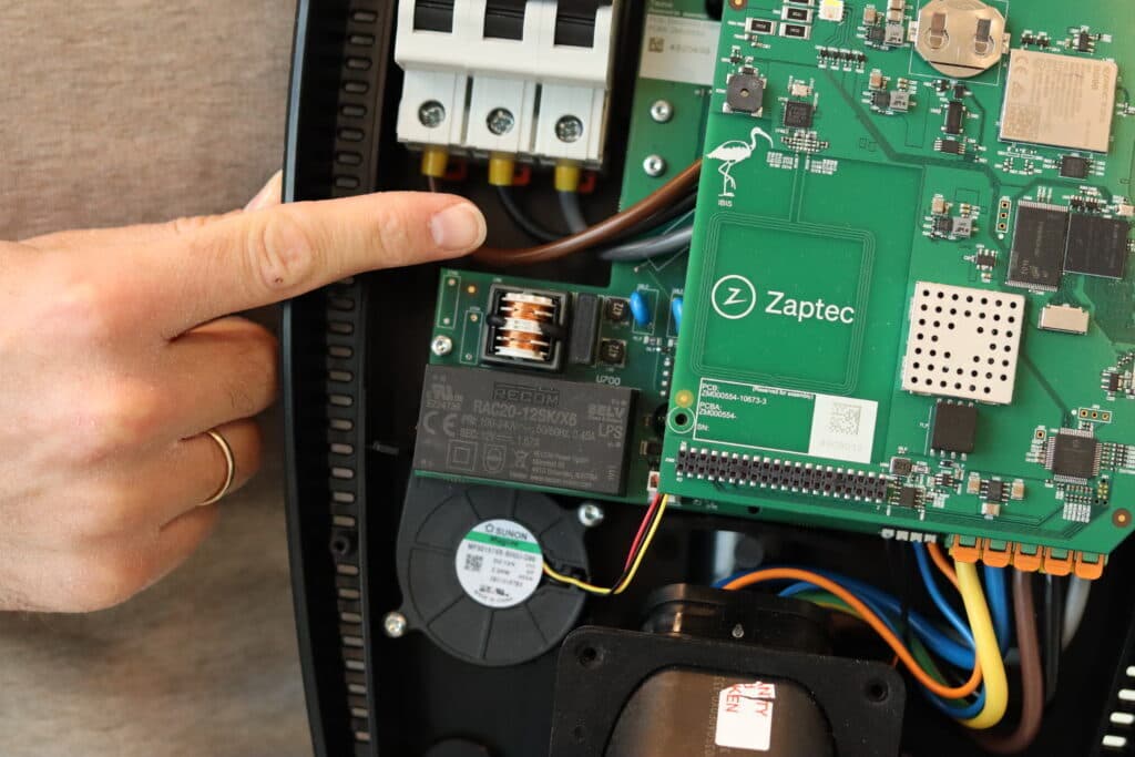 En person peker på en komponent inne i en åpen Zaptec-lader, der kretskort og elektriske systemer er tydelig synlige.