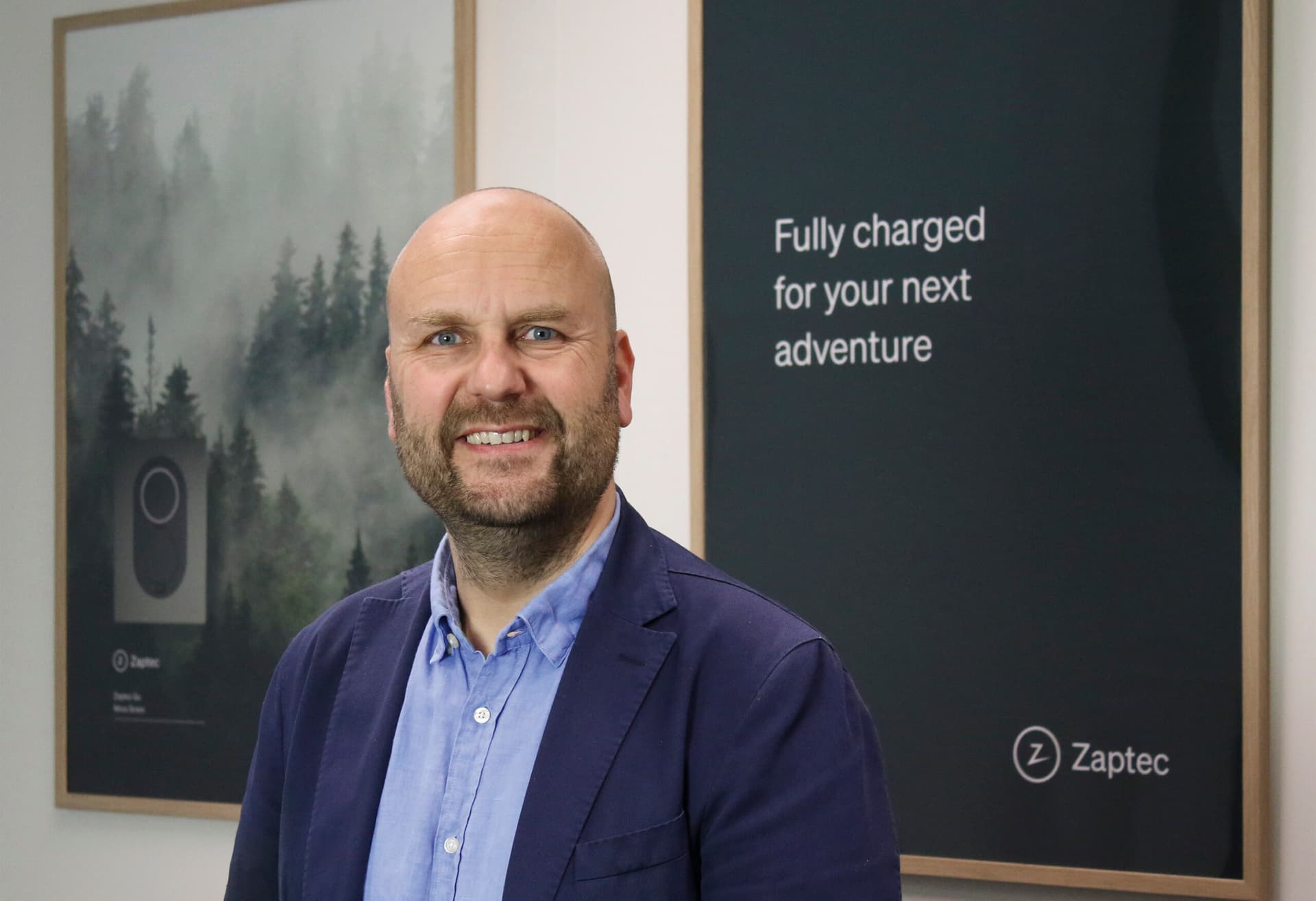Een glimlachende man in een blauwe blazer poseert voor Zaptec-promotiemateriaal. Op de achtergrond hangen posters, waarvan één met de tekst "Fully charged for your next adventure" en een afbeelding van een laadstation omringd door een boslandschap.