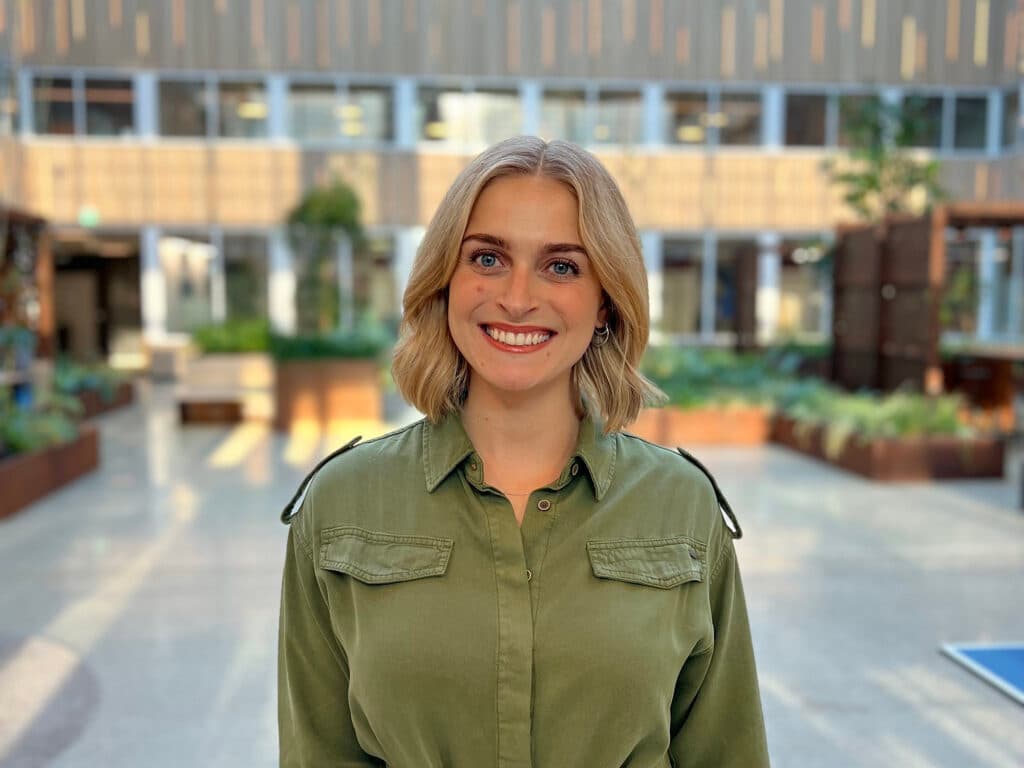 En smilende kvinne med blondt hår og grønn skjorte står i et lyst og moderne atrium med planter og store vinduer i bakgrunnen.