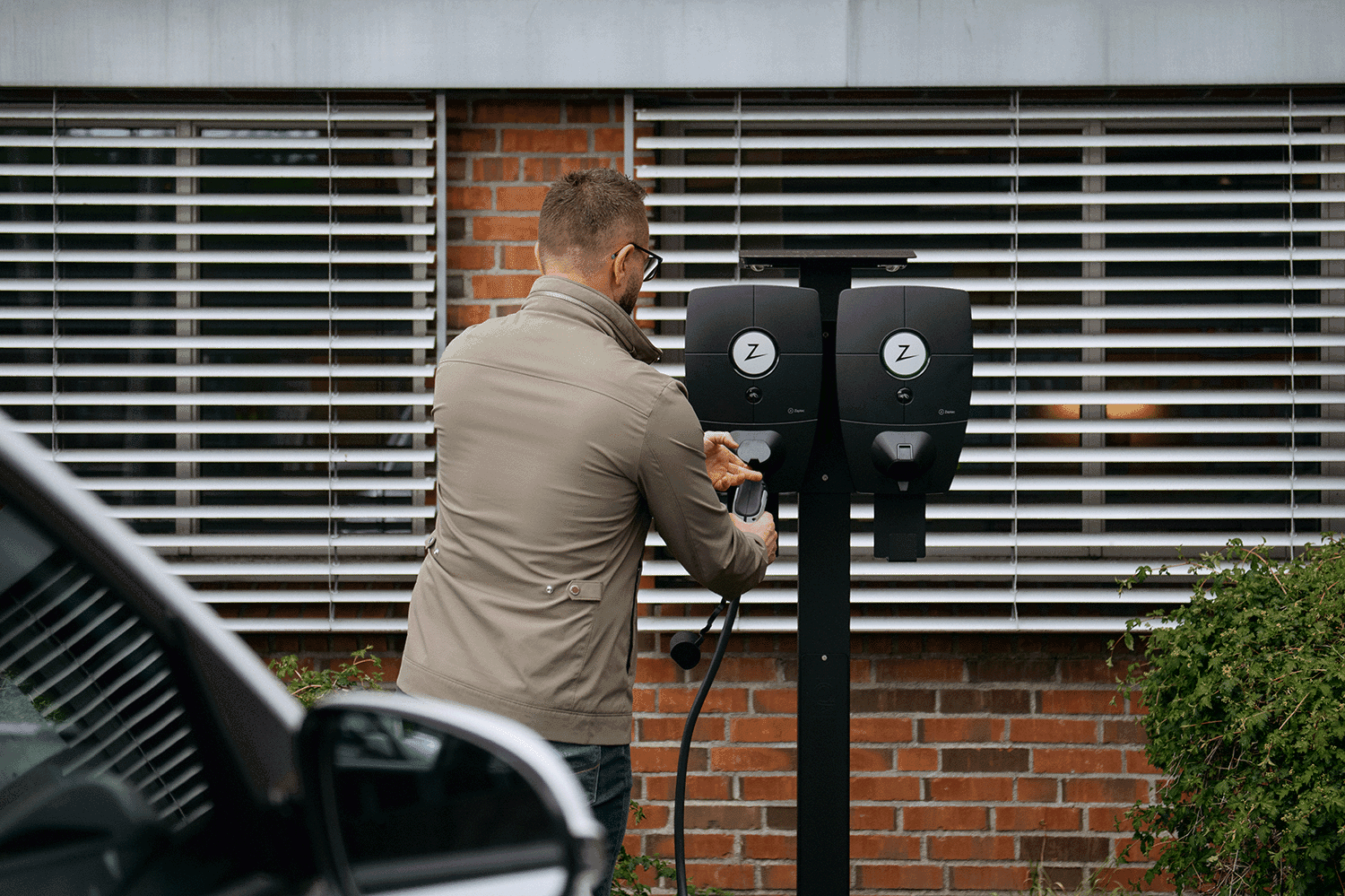 framför en tegelfasad sitter två Zapec Pro laddboxar monterad på ett stolpe en man i grön jacka kopplar in en laddsladd i den ena laddaren, i framkant skymtar man en vit elbil