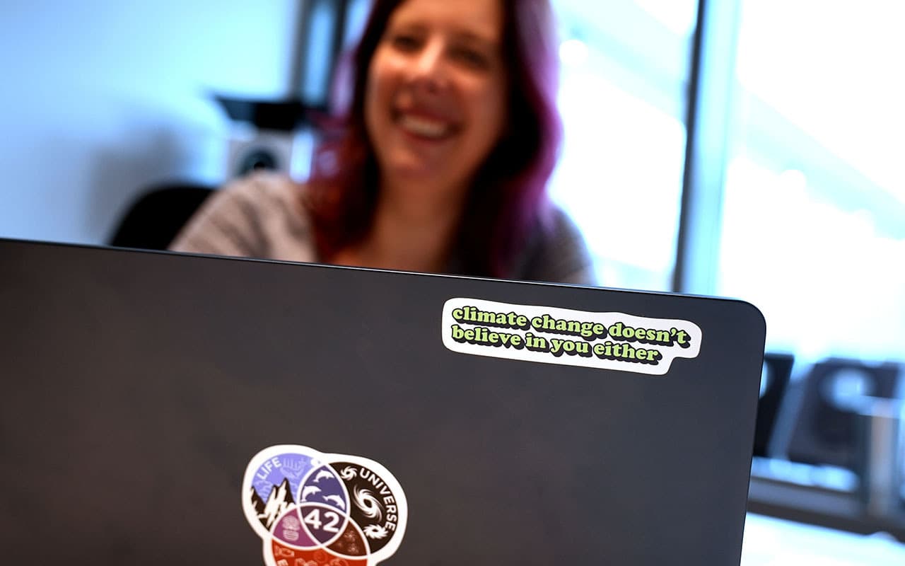 En kvinna med mörkt hår med röd-rosa toner och grå tröja syns bakom en svart laptop som står framför henne