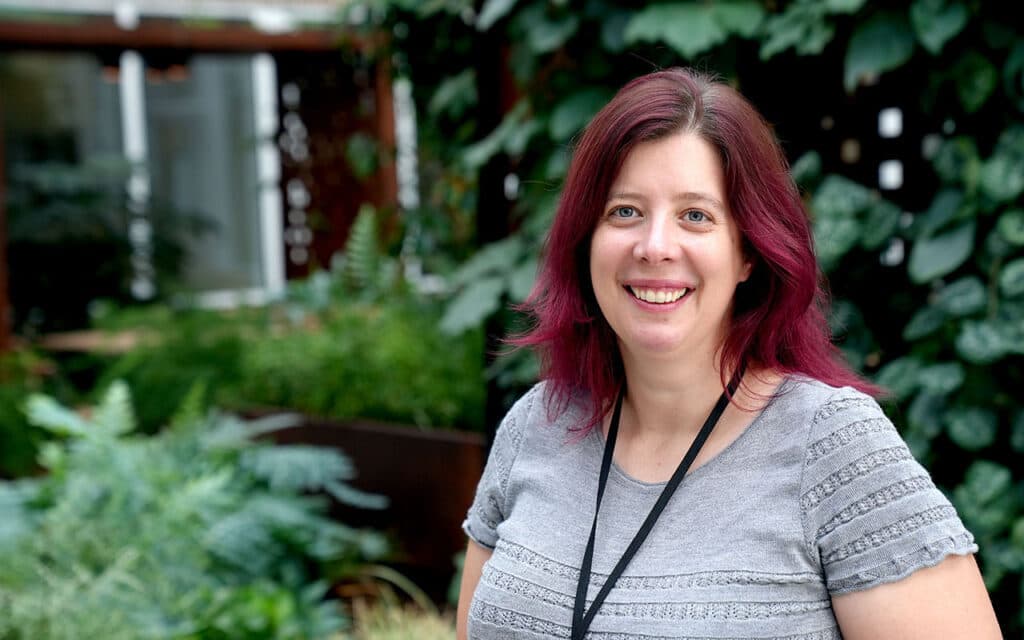 En kvinna med mörkt hår med röd-rosa toner och grå tröja kollar leende mot kameran. i bakgrunden är det gröna växter.