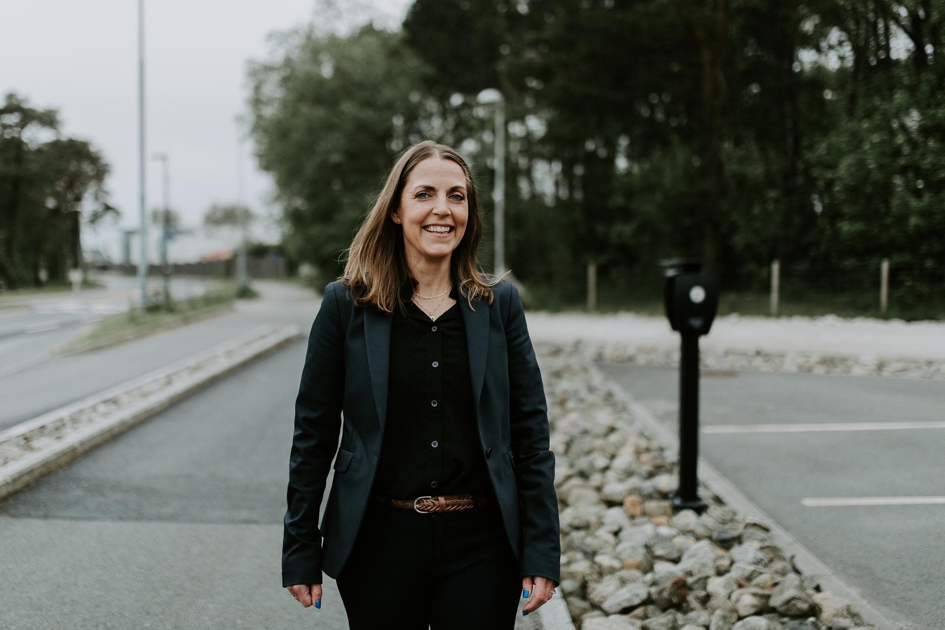 En kvinne i mørk blazer går smilende langs en vei med en Zaptec Pro-lader synlig i bakgrunnen ved en parkeringsplass.