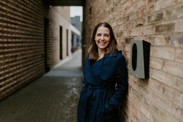 En kvinne i mørk blå kåpe står smilende lent mot en mursteinsvegg der en Rock Grey Zaptec Go-lader er montert.