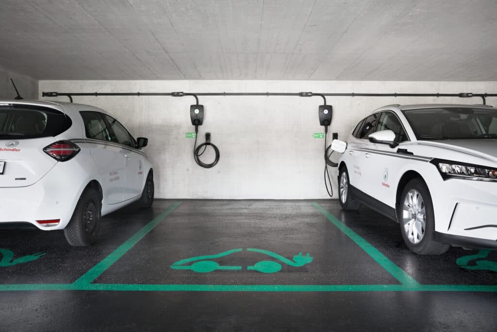 To hvite elbiler med Schindler-logo står parkert i et garasjeanlegg med dedikerte ladeplasser, hvor den ene bilen lades med en Zaptec Pro-lader montert på betongveggen.