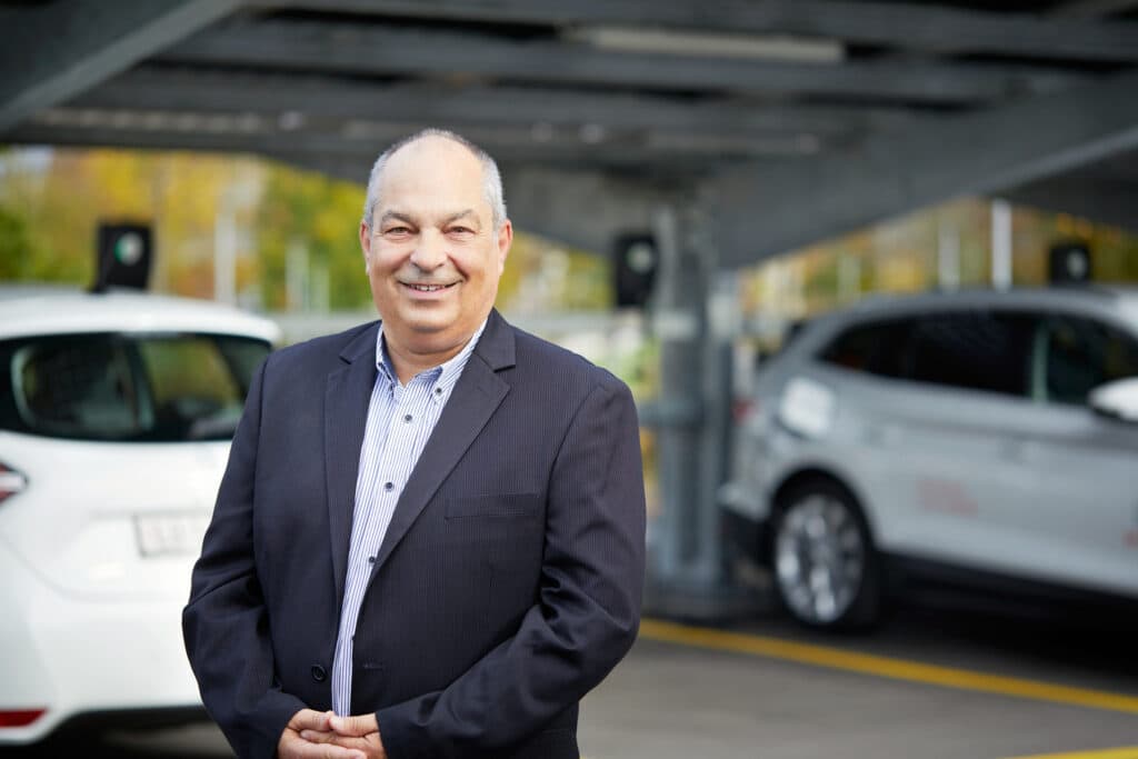 En mann i dress står smilende foran to hvite elbiler som lades med Zaptec Pro-ladere under et utendørs parkeringsskur.