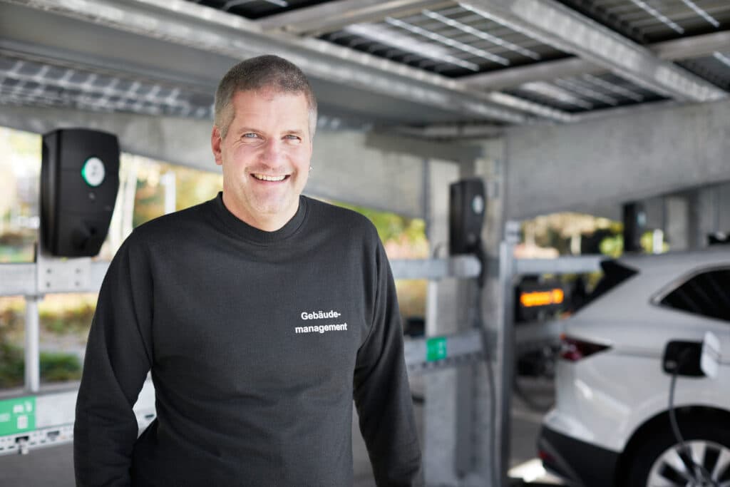 En mann med genser med teksten "Gebäudemanagement" står smilende foran ladende elbiler ved et parkeringsskur utstyrt med Zaptec Pro-ladere.