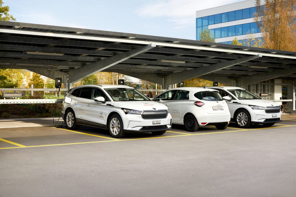 Tre hvite elbiler med Schindler-logo står parkert på rekke under et solcelledrevet parkeringsskur, hvor en av bilene lades med en Zaptec Pro-lader.