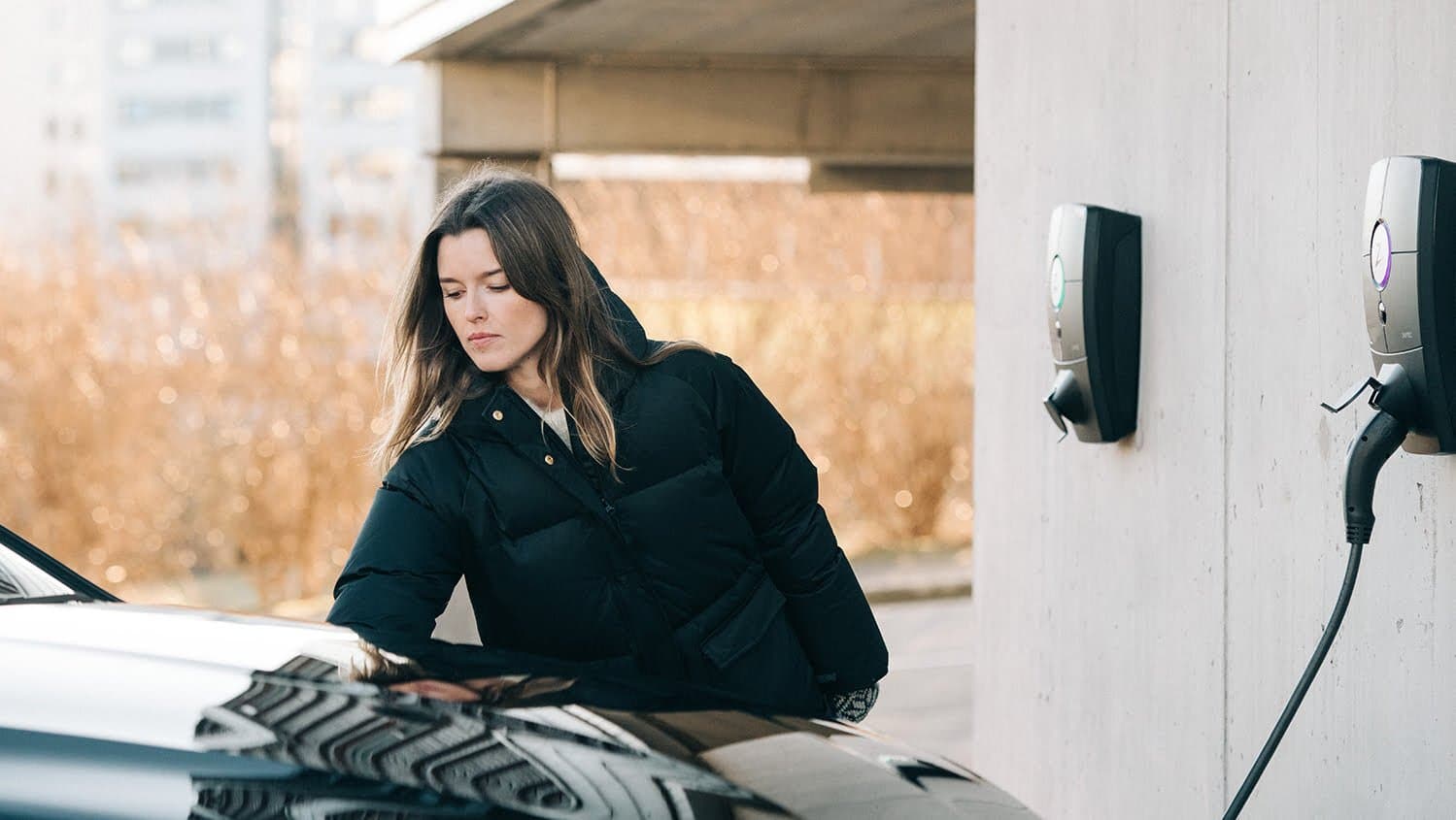 En kvinna i en svart vinterjacka står vid en bil som är ansluten till en Zaptec Pro laddstation monterad på en betongvägg.