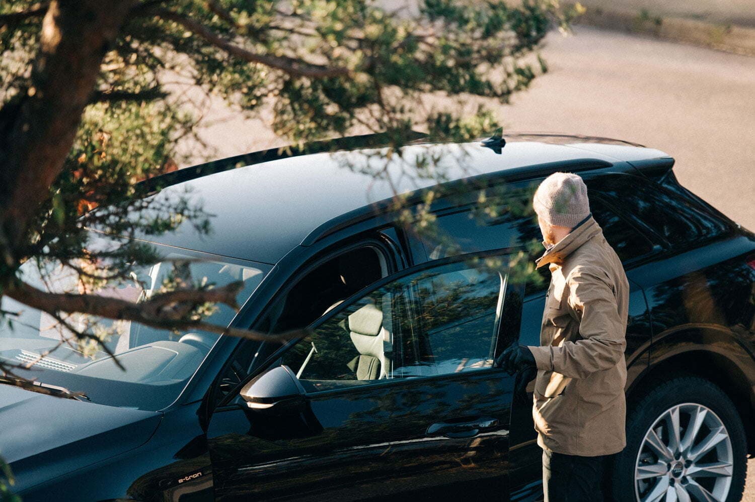 En mann med grå lue og beige jakke åpner døren til en svart elbil som står parkert i solen, delvis skjult av et tre.