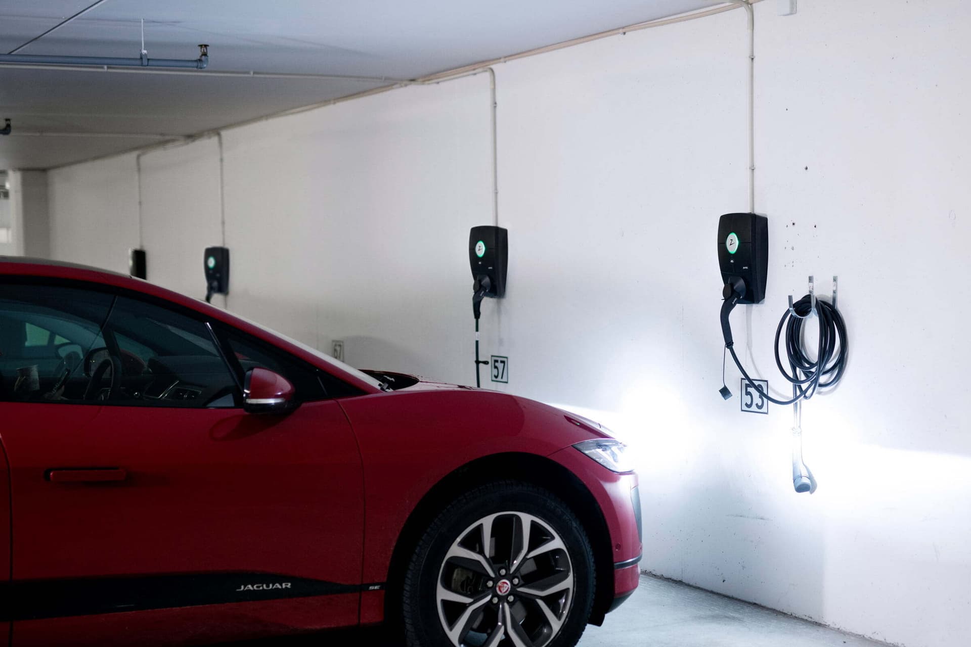 En rød elbil står parkert i en garasje foran en rekke Zaptec Pro-ladere montert på en hvit vegg. En av laderne er i bruk, med kabelen koblet til bilen.