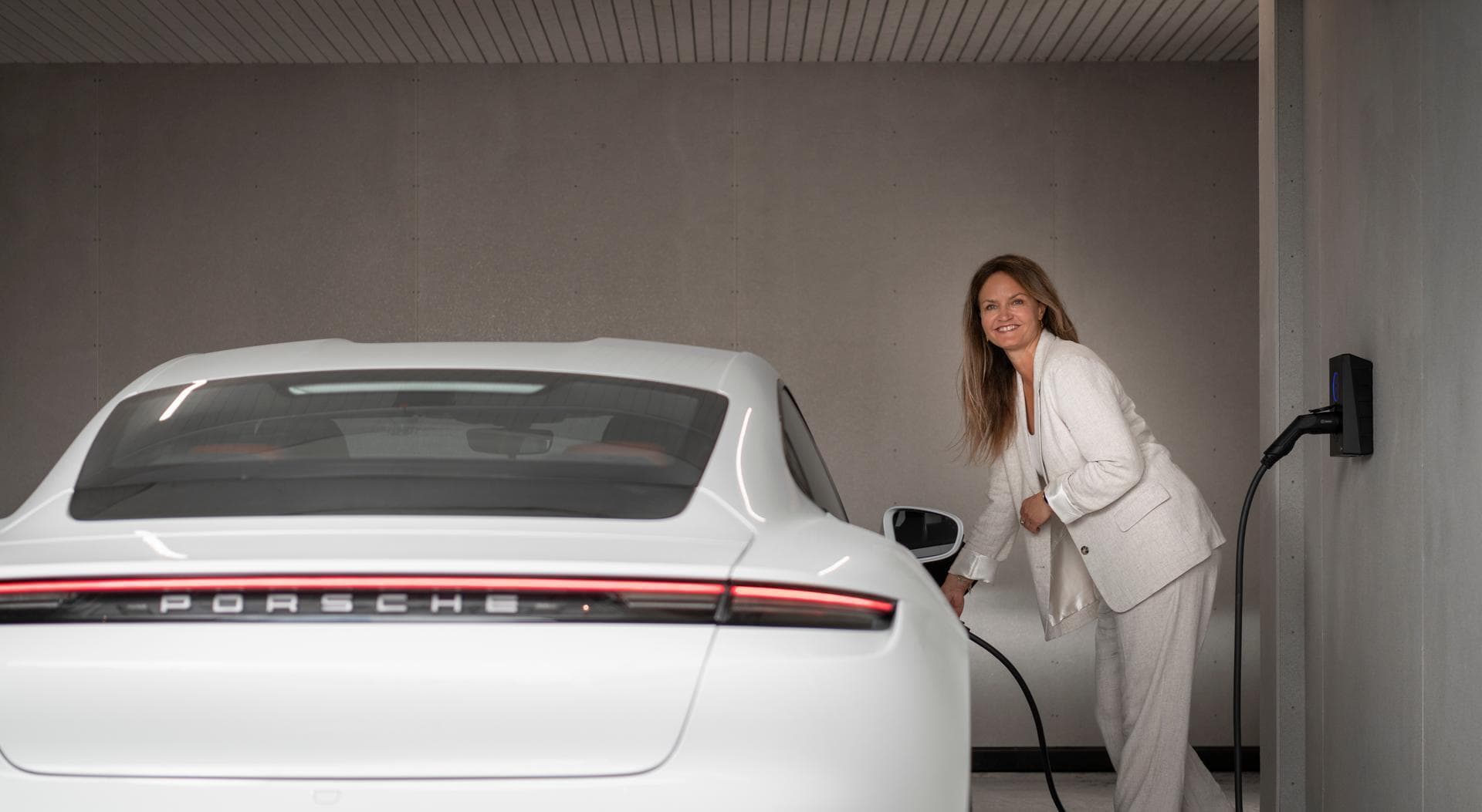 En kvinne i lys dress lader en hvit Porsche med en Asphalt Black Zaptec Go-lader montert på en lys vegg i en garasje.