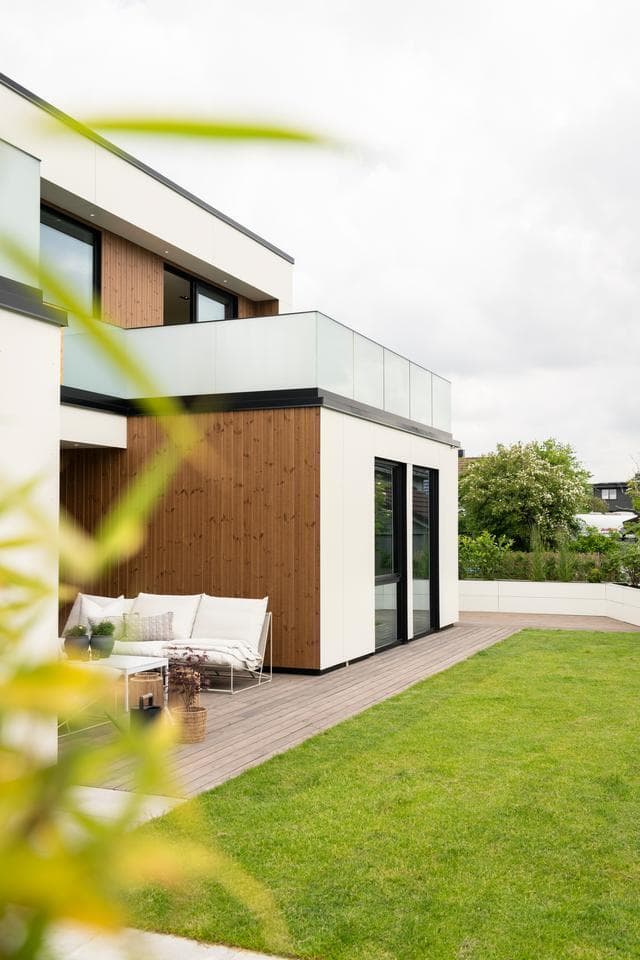 Et moderne hus med hvit fasade og trepanel har en koselig uteplass med sofa og frodig hage i forgrunnen.