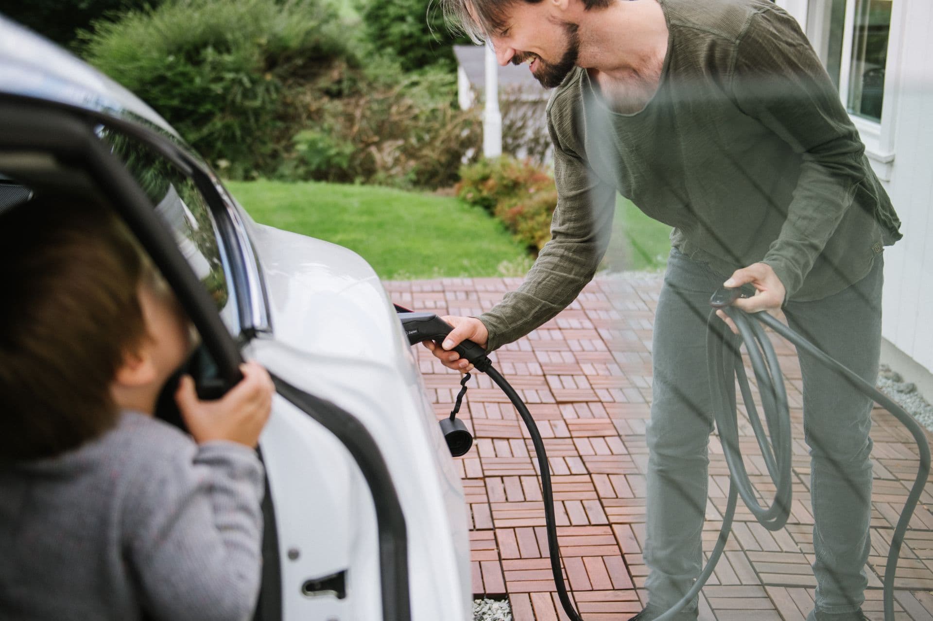 En mann holder en ladekabel og smiler til et barn som sitter i en hvit elbil, sett gjennom bilvinduet.