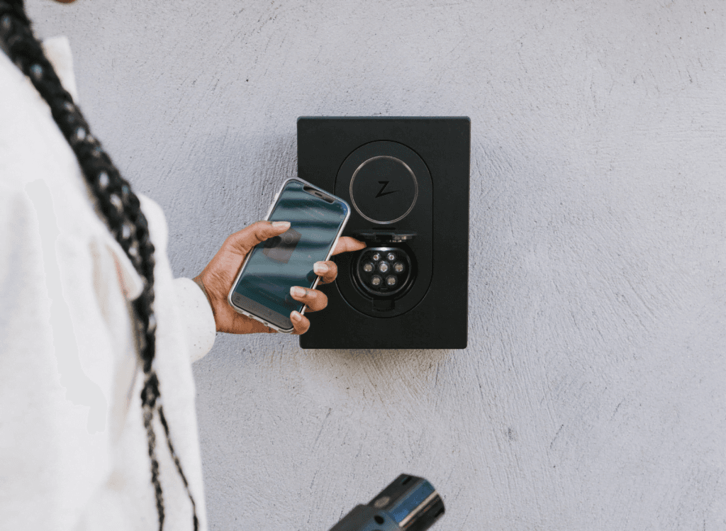 En person holder en mobiltelefon foran en Asphalt Black Zaptec Go-lader som er montert på en lys betongvegg. I den andre hånden holder vedkommende en ladekabel klar til bruk.
