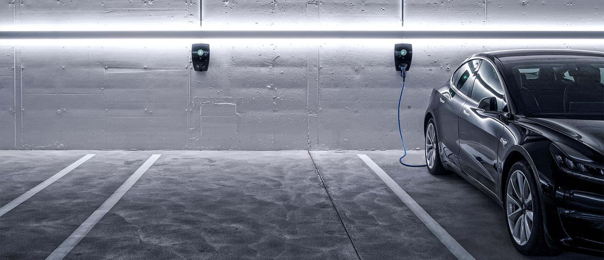 En svart elbil lades med en Zaptec Pro-lader i et moderne parkeringsanlegg med betongvegger og LED-lys langs taket. Flere ladeplasser er tilgjengelige ved siden av.