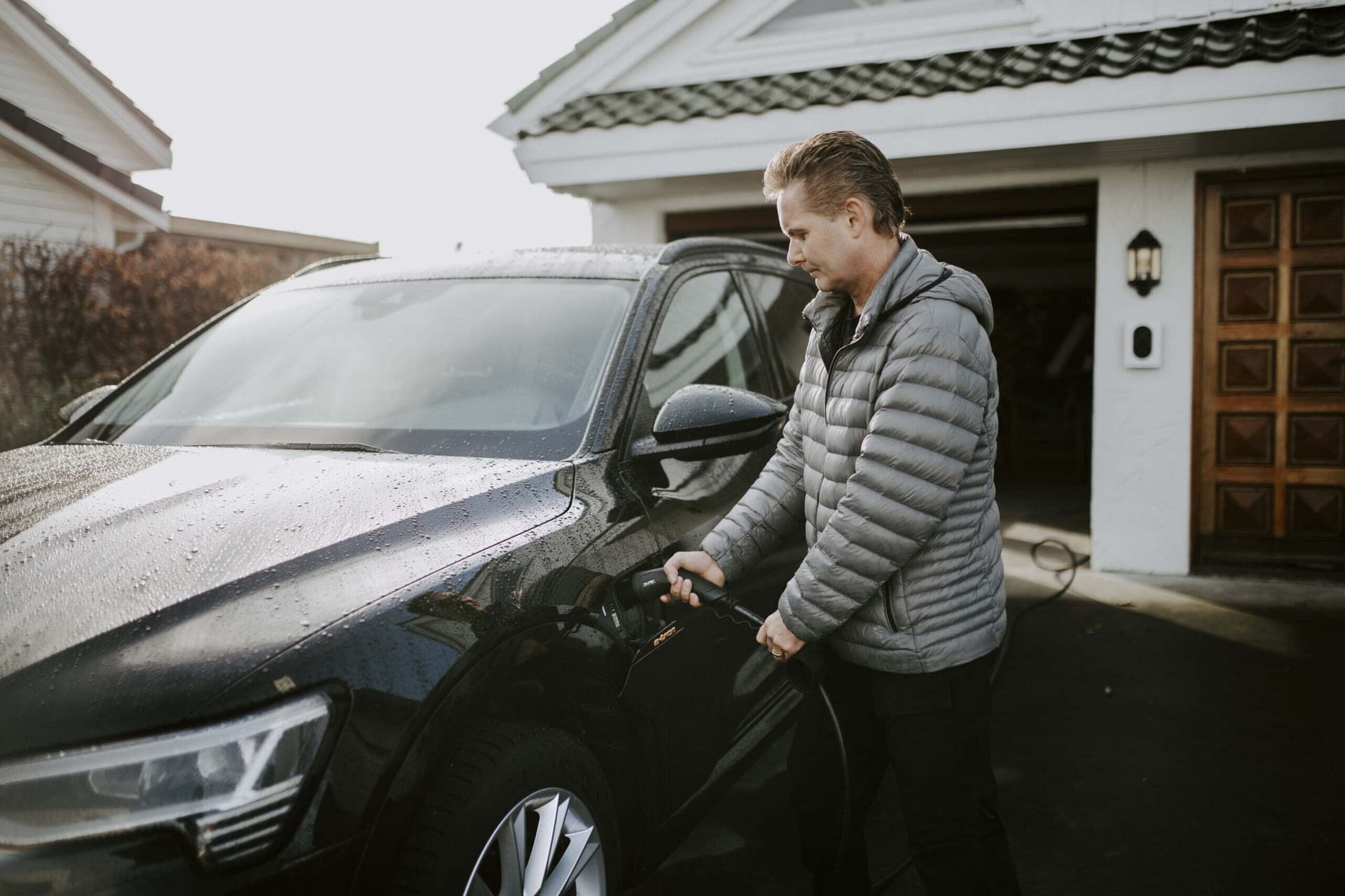 En mann i grå jakke lader en svart elbil foran garasjen hjemme. På veggen ved siden av garasjeporten er en hvit Zaptec Go-lader montert.