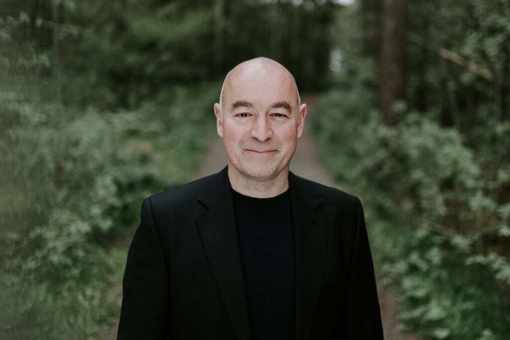 en man utan hår och mörk kavaj står i en grönskande miljö