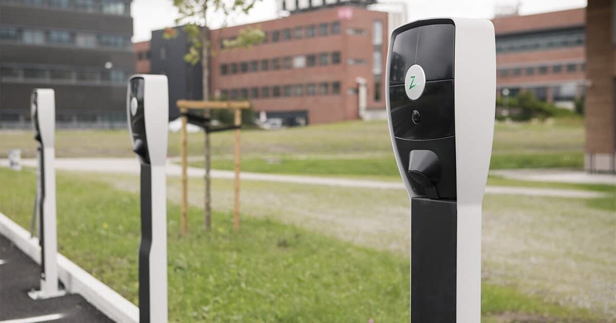 Flere Zaptec Pro-ladere med hvite sidepaneler står montert på rekke ved en parkeringsplass i et grønt område utenfor moderne kontorbygg.