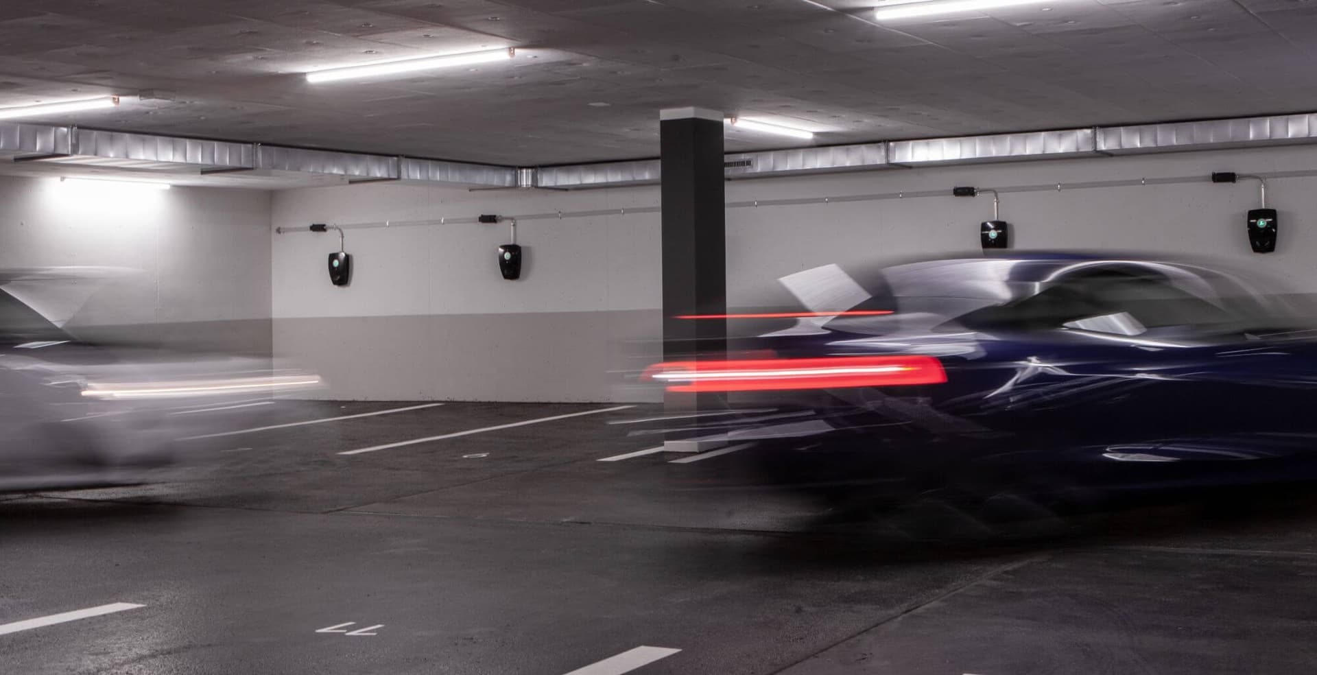To biler kjører gjennom en parkeringskjeller med flere Zaptec Pro-ladere montert på veggen. Bevegelsesuskarphet viser farten til bilene i det moderne, godt opplyste anlegget.