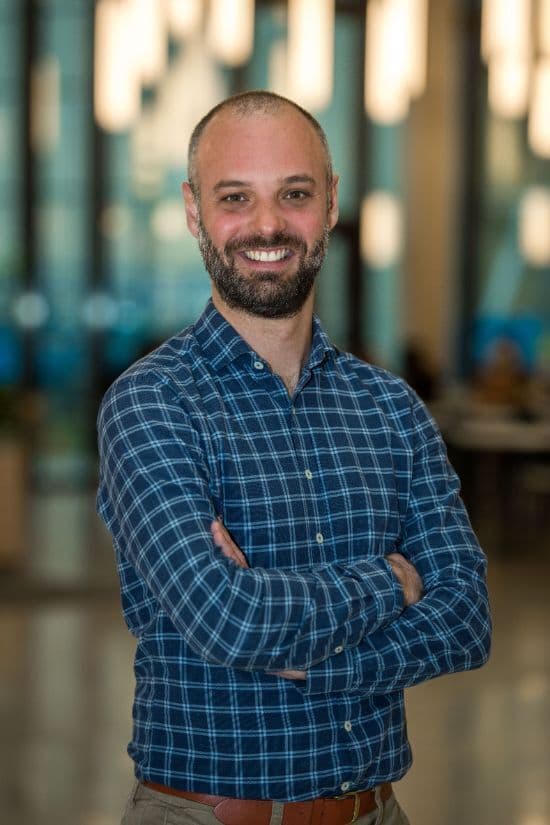 Smilende mann med skjegg og rutete blå skjorte står med armene i kors innendørs i et lyst kontormiljø