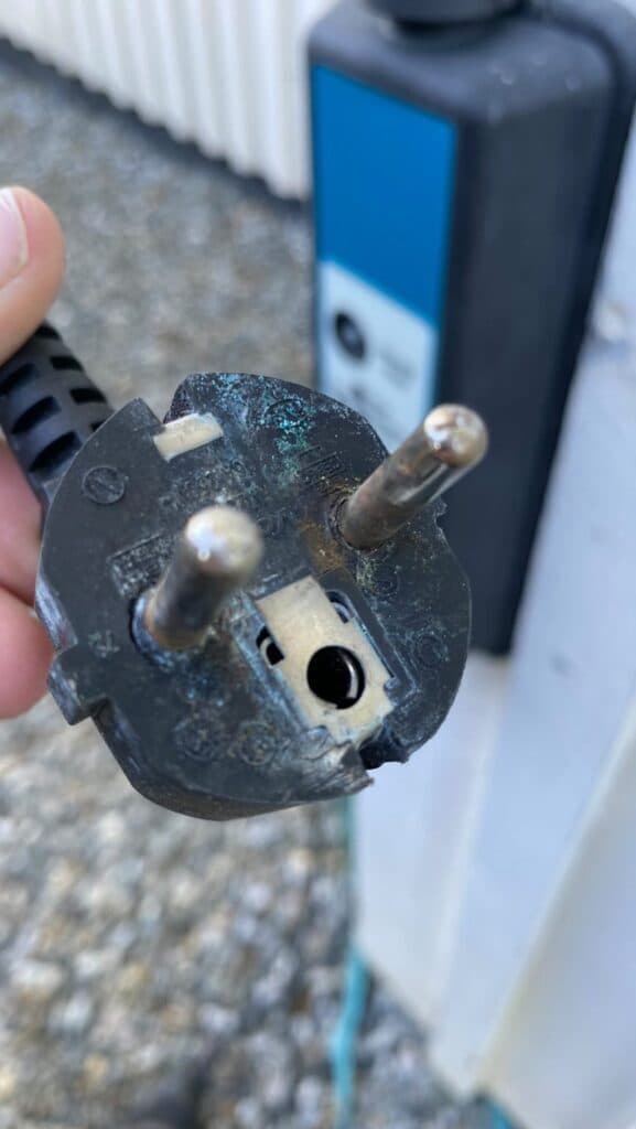 Nærbilde av en svart, skadet støpsel med synlige brennmerker og irr på metallpinnene. Støpselet holdes i hånden foran en hvit vegg og en blå ladestasjon i bakgrunnen