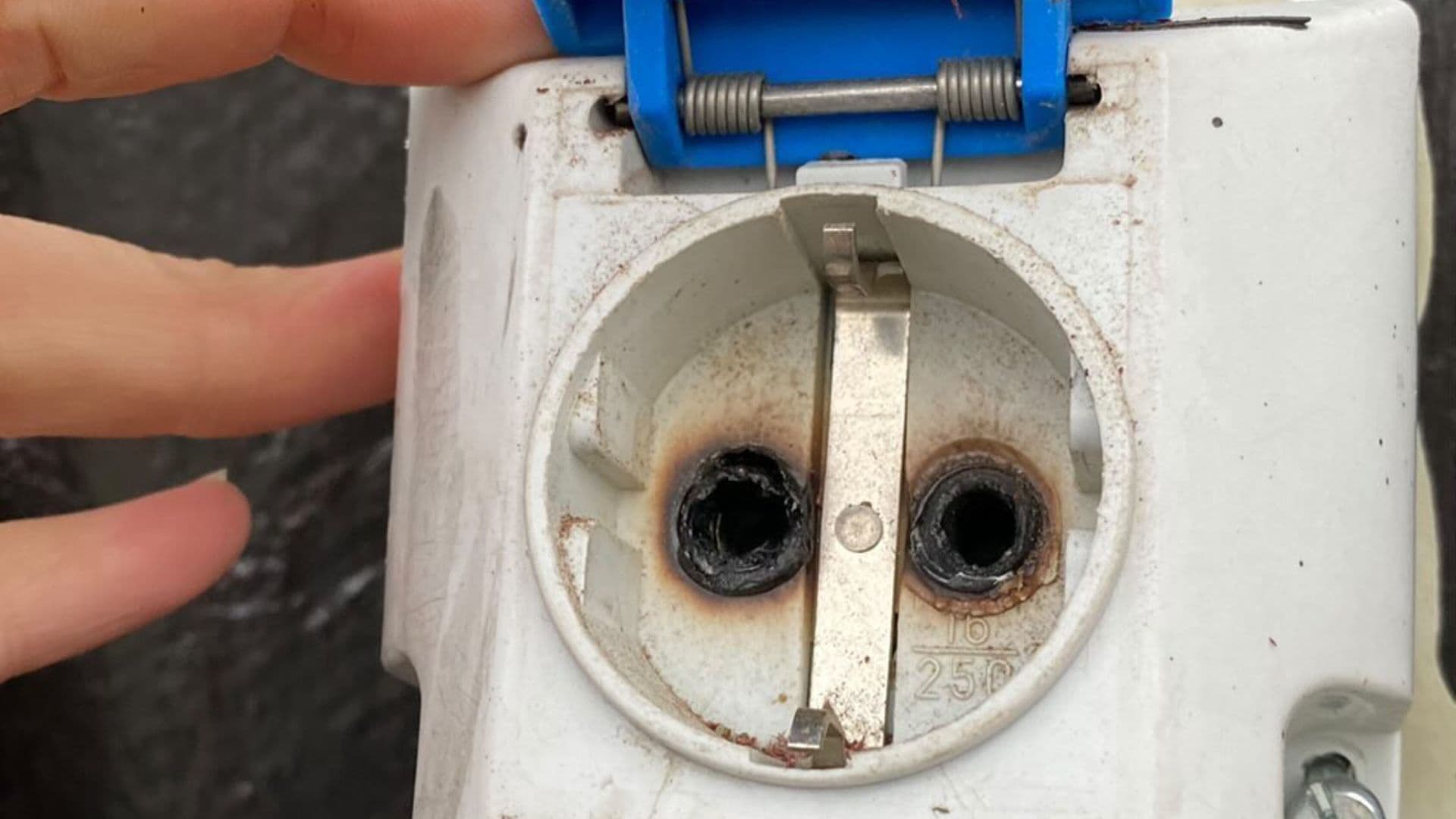 Nærbilde av en brent og skadet stikkontakt med svidde kontaktpunkter og misfarging rundt hullene. En hånd holder lokket åpent for å vise skadene.