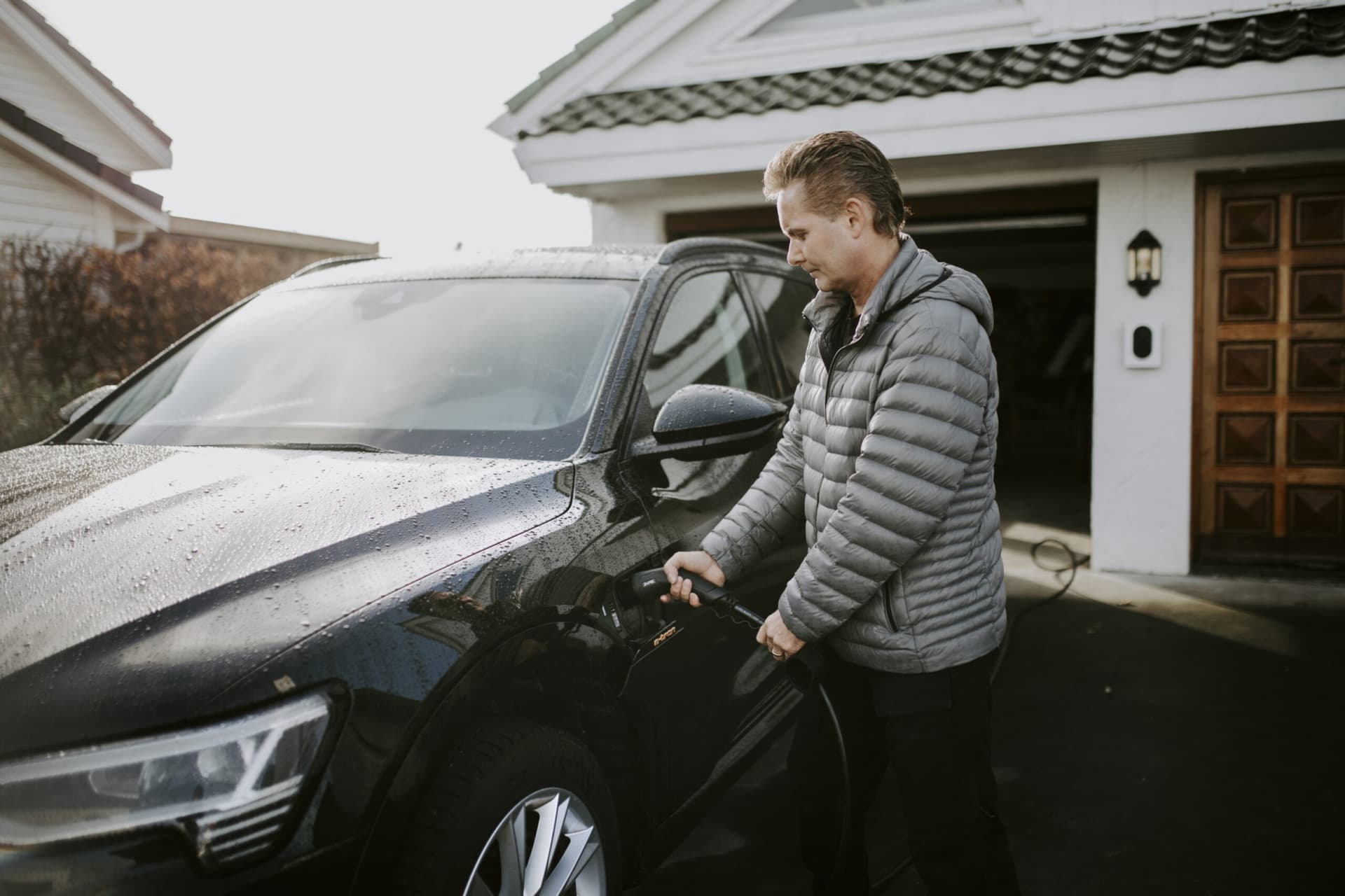 En man med grå jacka kopplar in en laddkabel i sin svarta elbil, i bakgrunden sitter en vit Zaptec Go monterad på den vita fasaden mellan två mörka dörrar.