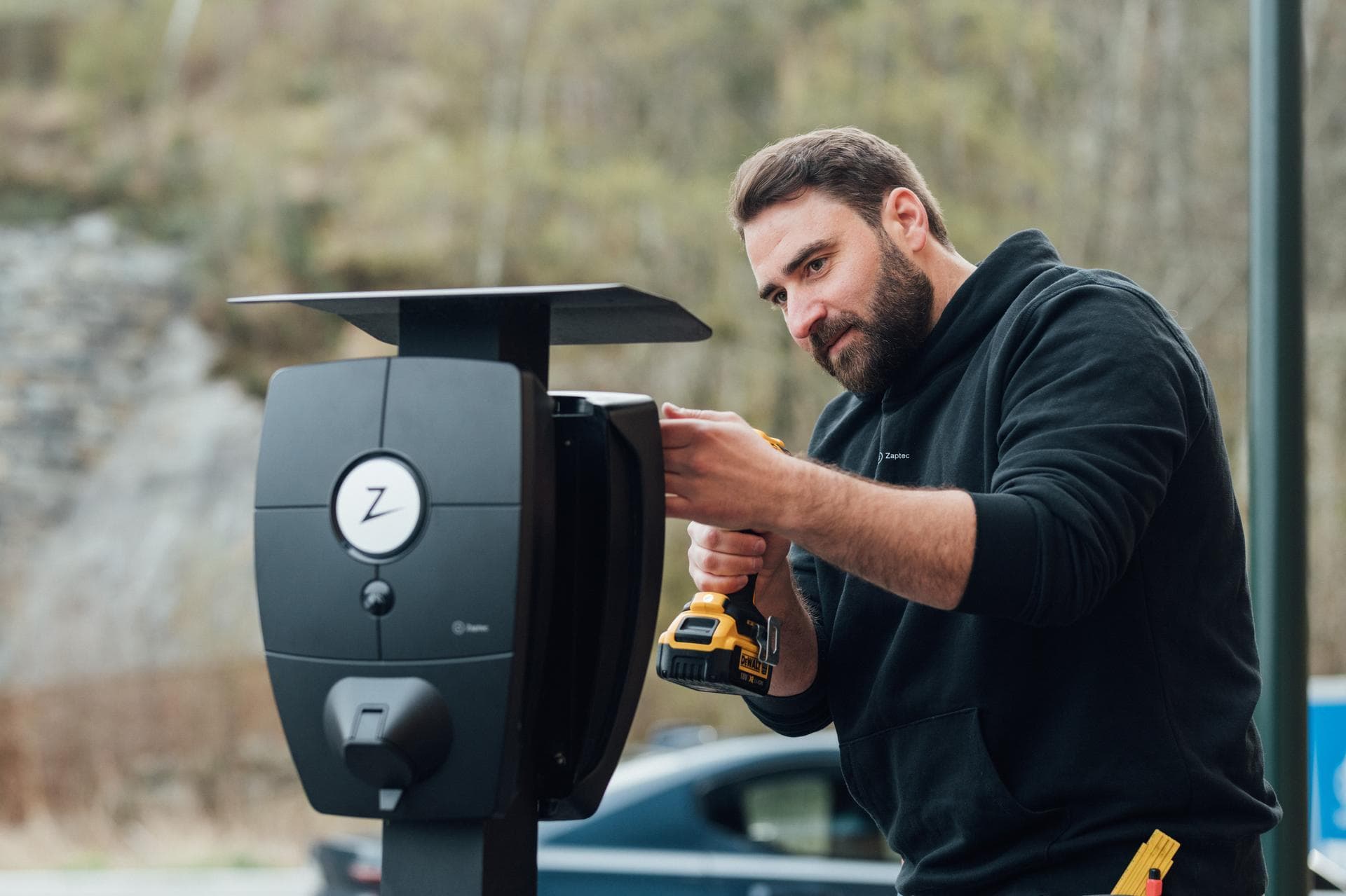 En man i en svart hoodie installerar en Zaptec Pro laddbox för elbilar utomhus med hjälp av en borrmaskin. I bakgrunden syns träd och en stenig terräng.