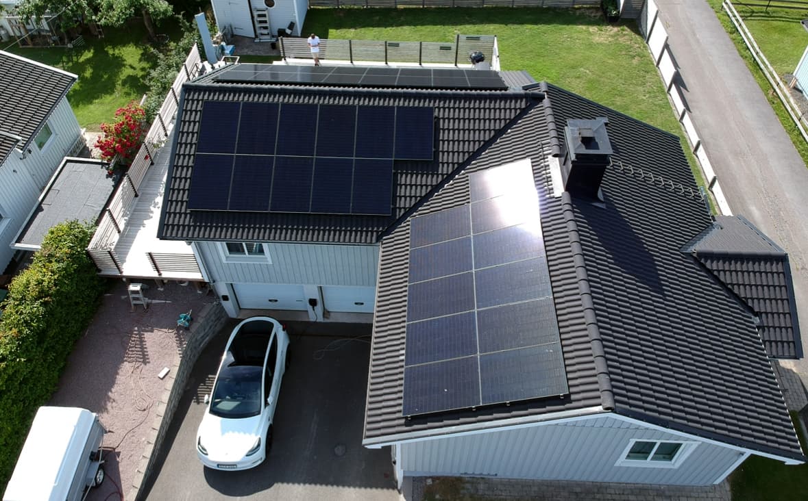 Ett ljusgråblått hus med svart tegeltak utrustat med stora solpaneler. En vit elbil står parkerad i uppfarten nedanför.