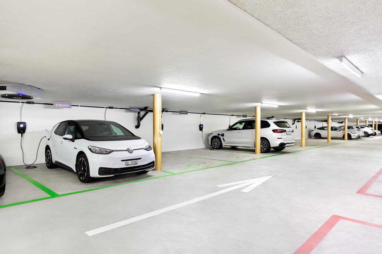 Flere hvite elbiler står parkert i et lyst parkeringsanlegg og lader med Zaptec Pro-ladere som er montert på veggen ved hver parkeringsplass.