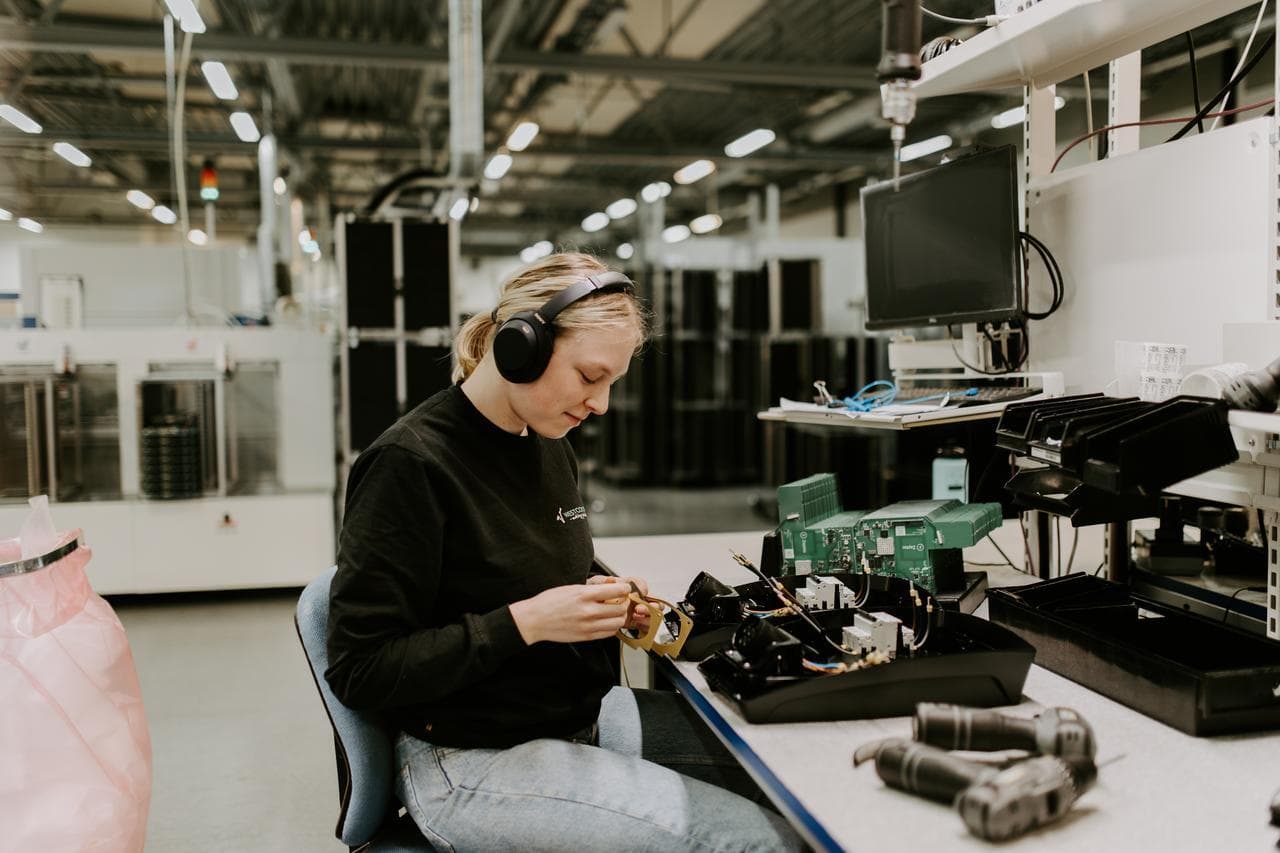 Kvinne med hodetelefoner monterer en Zaptec Pro elbillader ved en arbeidsbenk i et moderne produksjonslokale, med verktøy og dataskjerm på bordet.