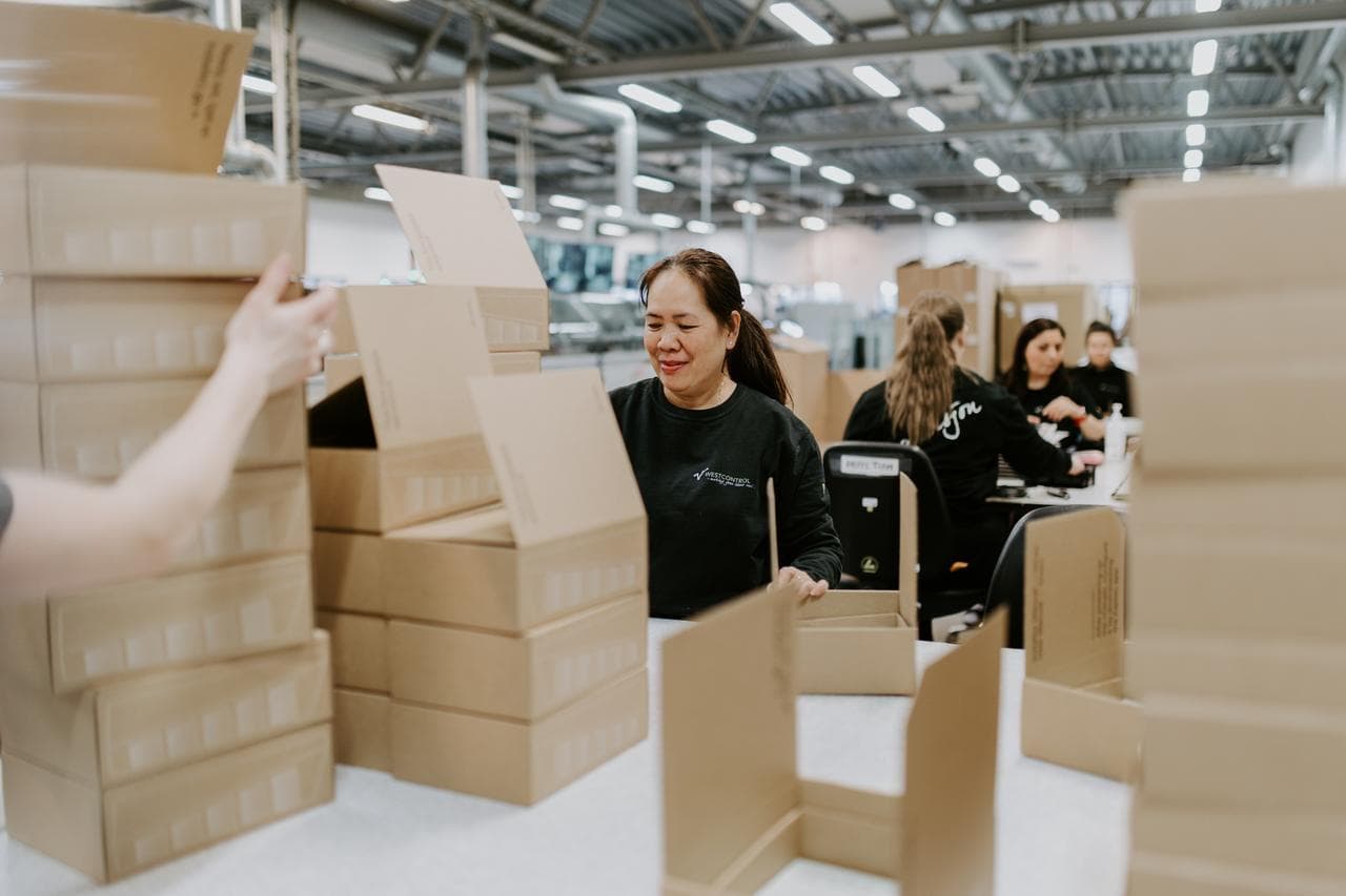 Flera personer sitter vid arbetsbänkar och packar Zaptec Go laddare i kartonger. På de vita bänkarna står travar av wellpapp kartonger med vitt tryck på som föreställer sidan av en laddbox.