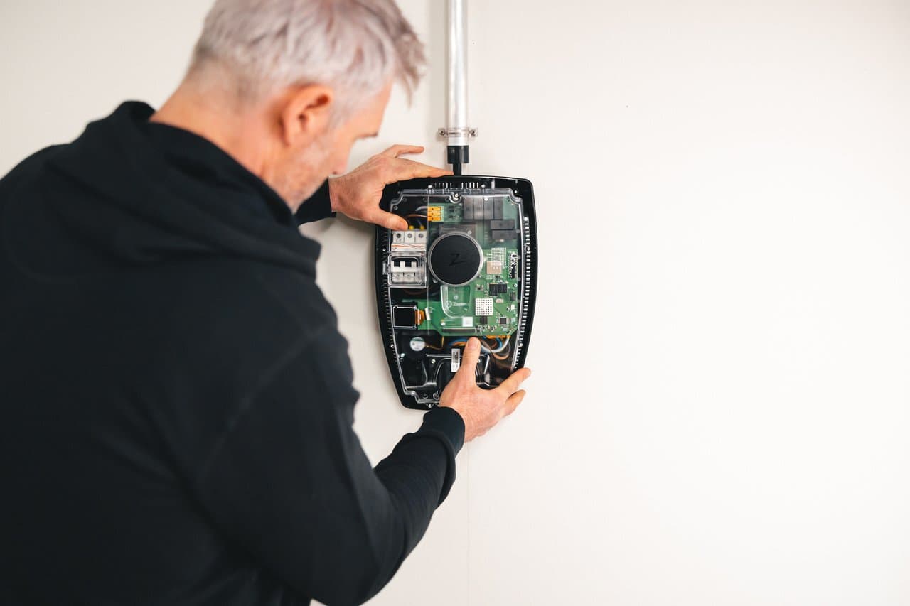 En mann i svart hettegenser justerer posisjonen til en Zaptec Pro ladestasjon for elbiler før montering på veggen i et lyst parkeringshus.