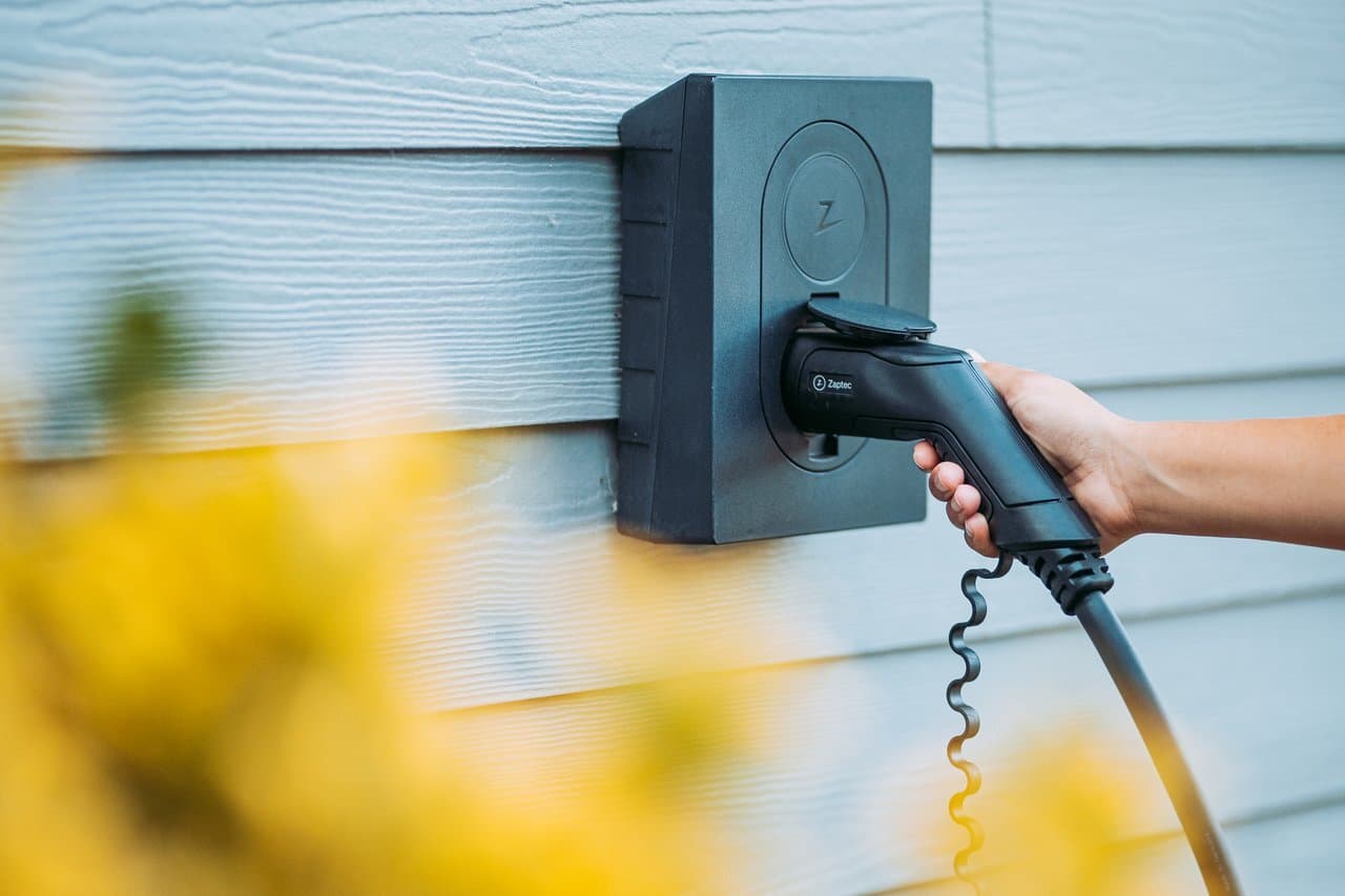 En hånd plugger inn en ladekabel i en Asphalt Black Zaptec Go-lader montert på en lys grå trevegg. Gule blomster er uskarpe i forgrunnen.