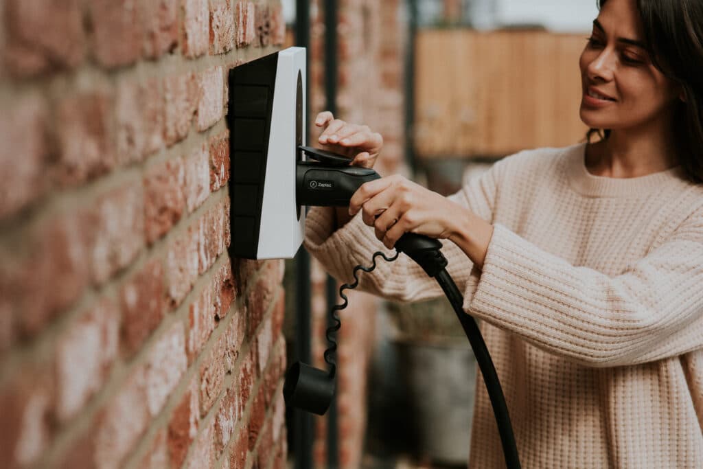 En kvinne i lys strikkegenser kobler til en ladekabel i en Cloud White Zaptec Go-lader som er montert på en rød mursteinsvegg.