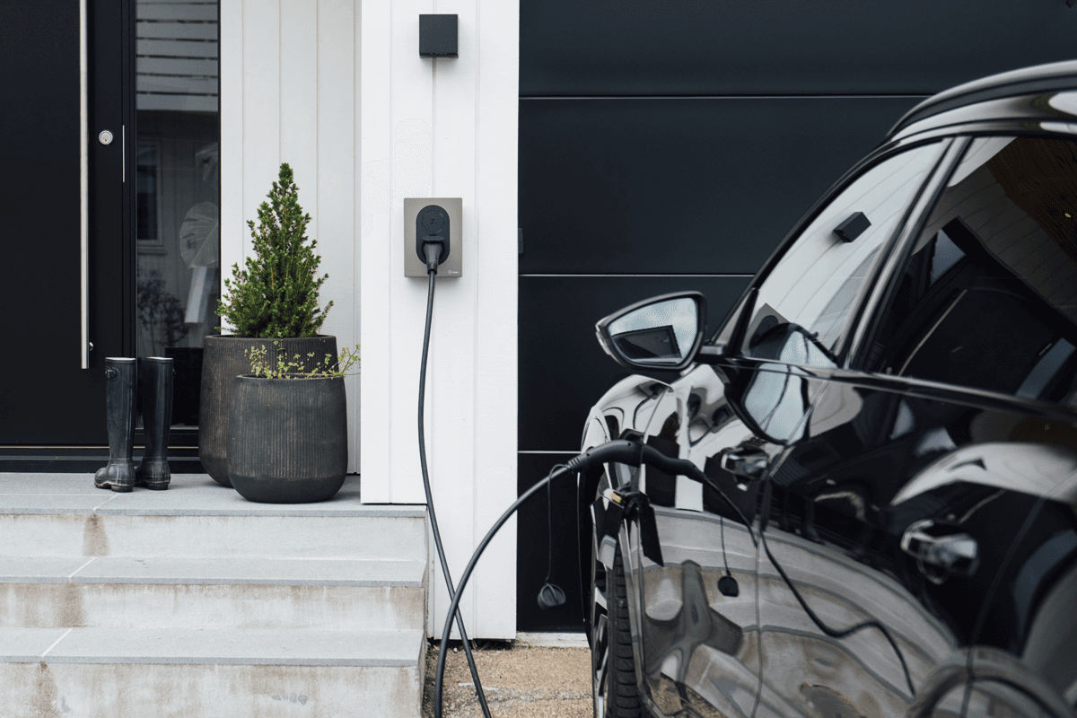 En svart bil lades med en Moss Green Zaptec Go-lader som er montert på en hvit trefasade. Ved døren står to mørke blomsterpotter med planter og et par svarte gummistøvler.