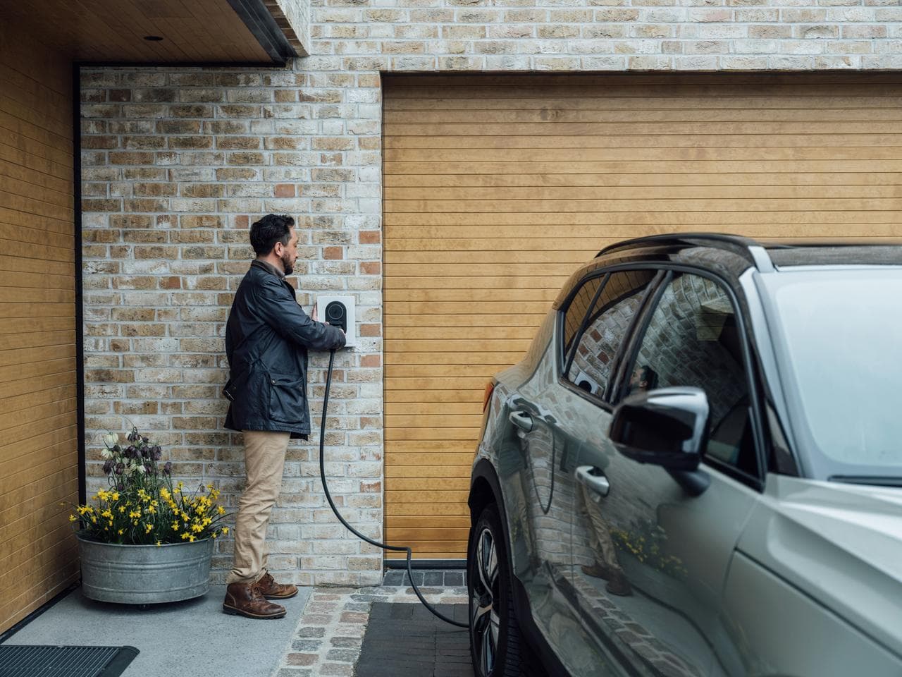 En mann i mørk frakk kobler til en Cloud White Zaptec Go-lader som er montert på en beige mursteinsvegg ved siden av en garasjeport i tre. En grønn-grå elbil står parkert foran laderen.