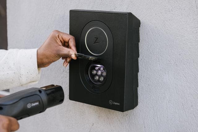 En person i en vit tröja öppnar ladduttaget på en svart Zaptec Go laddare monterad på en ljusgrå vägg och håller i en laddkabel med den andra handen.