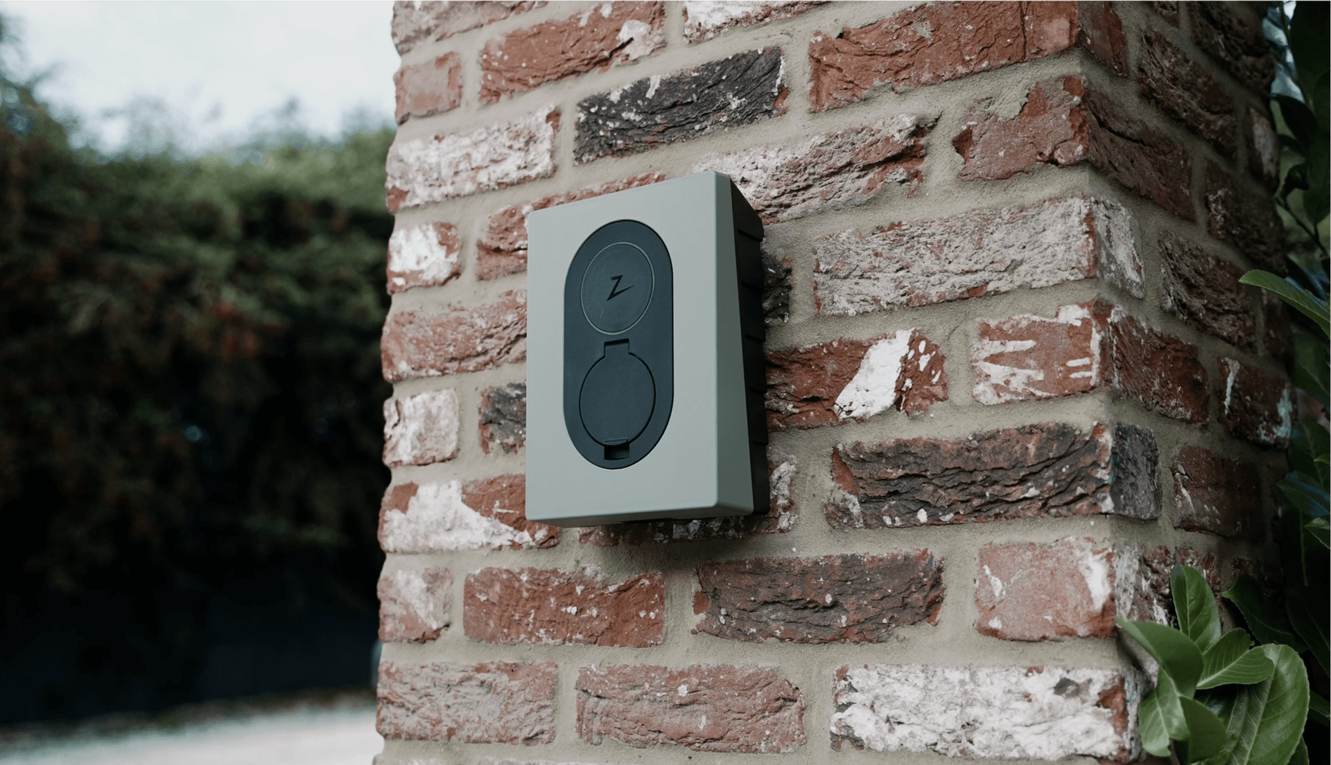 Grön Zaptec Go laddare sitter på en brun beige tegelvägg med gröna växter runtomkring.