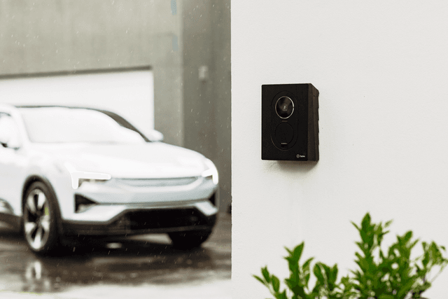 En Asphalt Black Zaptec Go 2 laddare är monterad på en vit vägg utanför ett modernt hem. I bakgrunden står en vit elbil parkerad på en regnblöt uppfart.